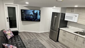 A modern living room with a black leather sofa, a flat-screen TV, a refrigerator, and a kitchen area with a microwave and sink. - Cozy Private 1-bedroom Basement Suite with free parking (Winnipeg)