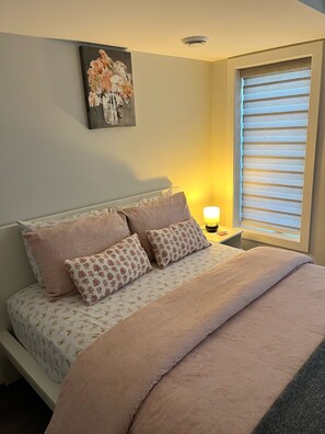 Una cama bien hecha con almohadillas estampadas con motivos florales y una manta color beige, una lámpara de noche y una obra de arte colgada en la pared sobre la cama.