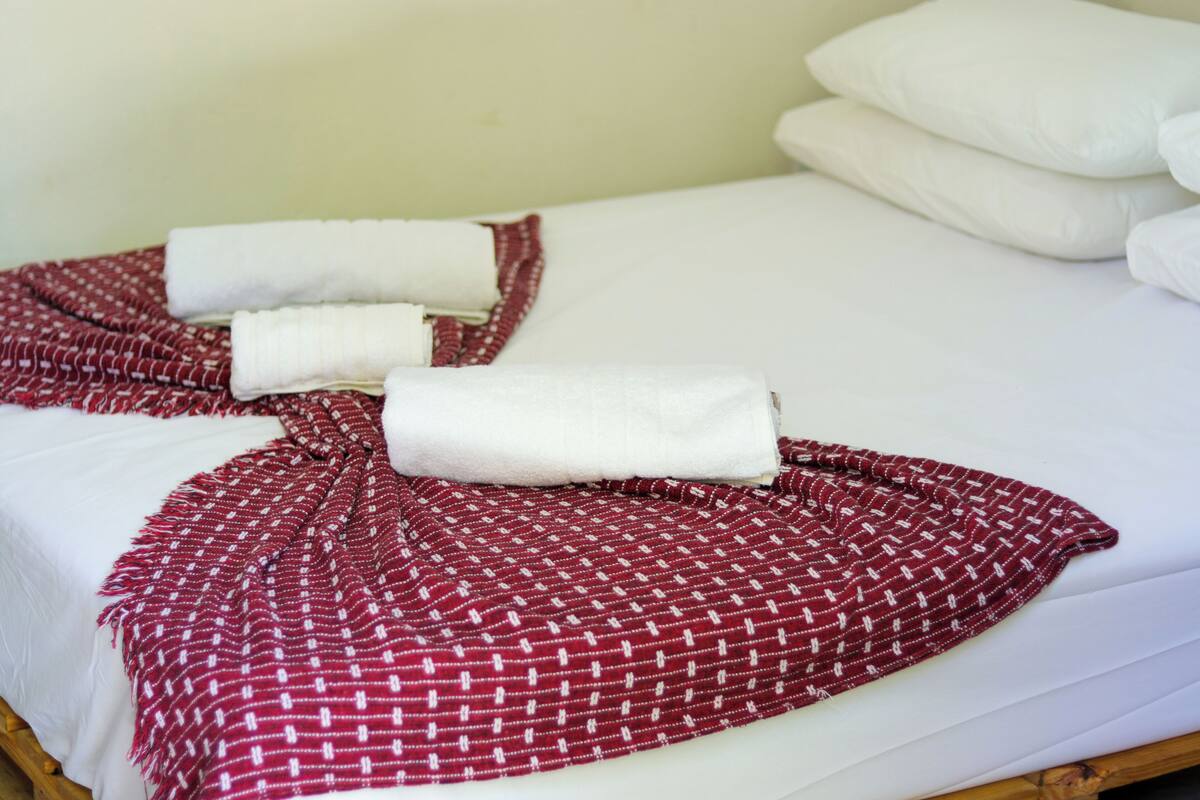 A bed with white linens, a red patterned blanket, and rolled towels.