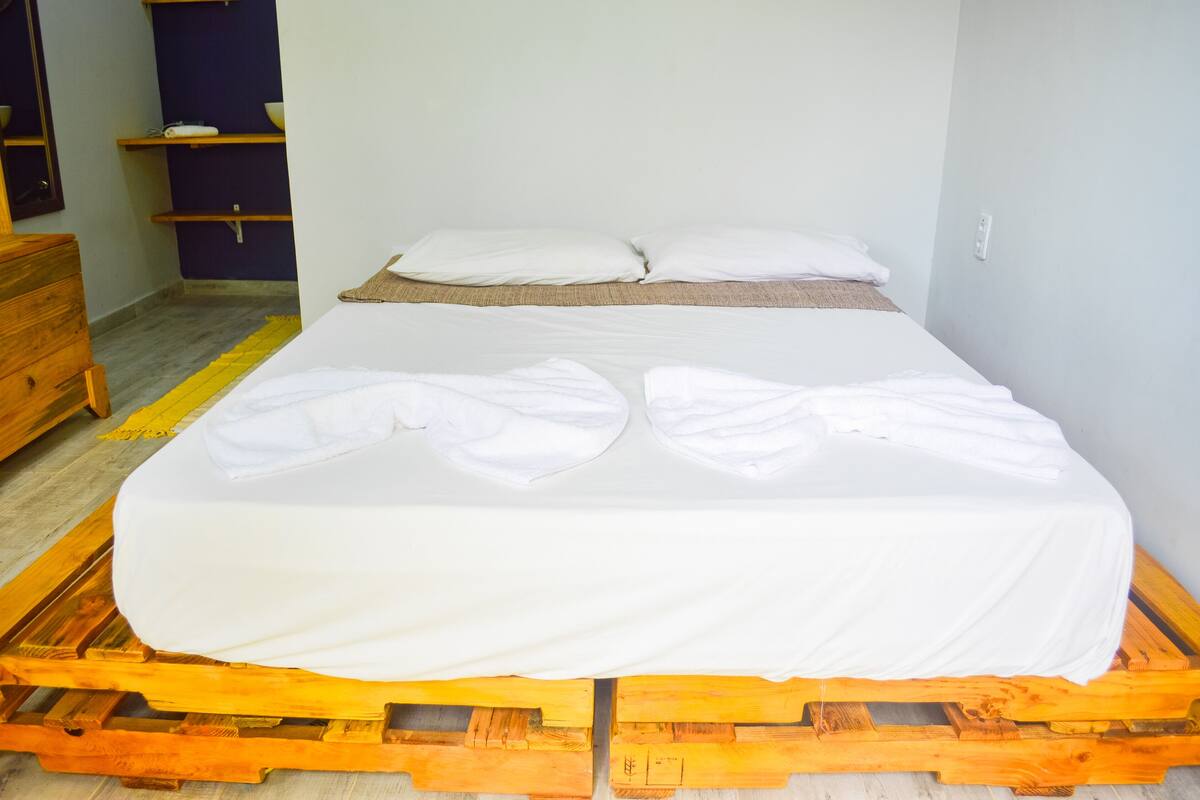 A single bed on a wooden pallet with white bedding, a wooden dresser, and a shelf unit with bowls.