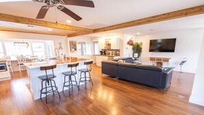 A spacious living area with a wooden floor, a dark sofa, a kitchen with white cabinets, and a large TV mounted on the wall. - Gulf Access Resort Style Pool Home Only 4 minutes to the Beach (Bonita Springs)