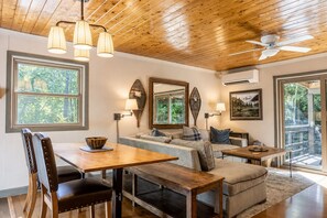 A spacious living area with a wooden ceiling, a dining table with chairs, a sofa, and a coffee table. There are large windows providing natural light and views of the outdoors.