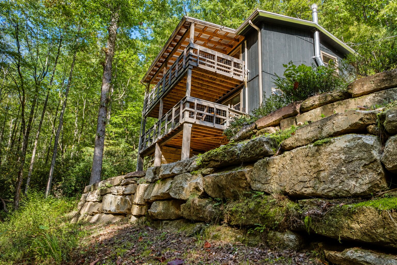 A cabin with a wooden deck and a stone retaining wall, surrounded by forest.