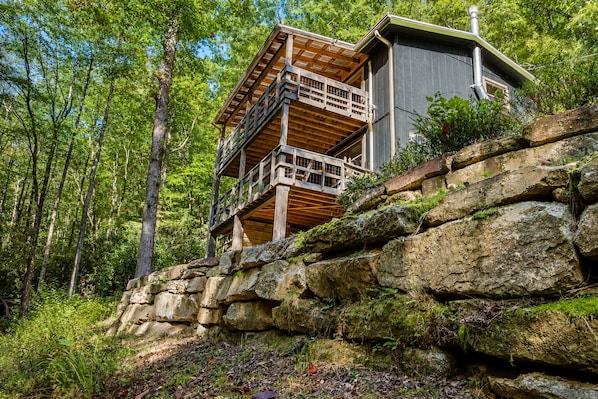 A cabin with a wooden deck and a stone retaining wall, surrounded by forest.