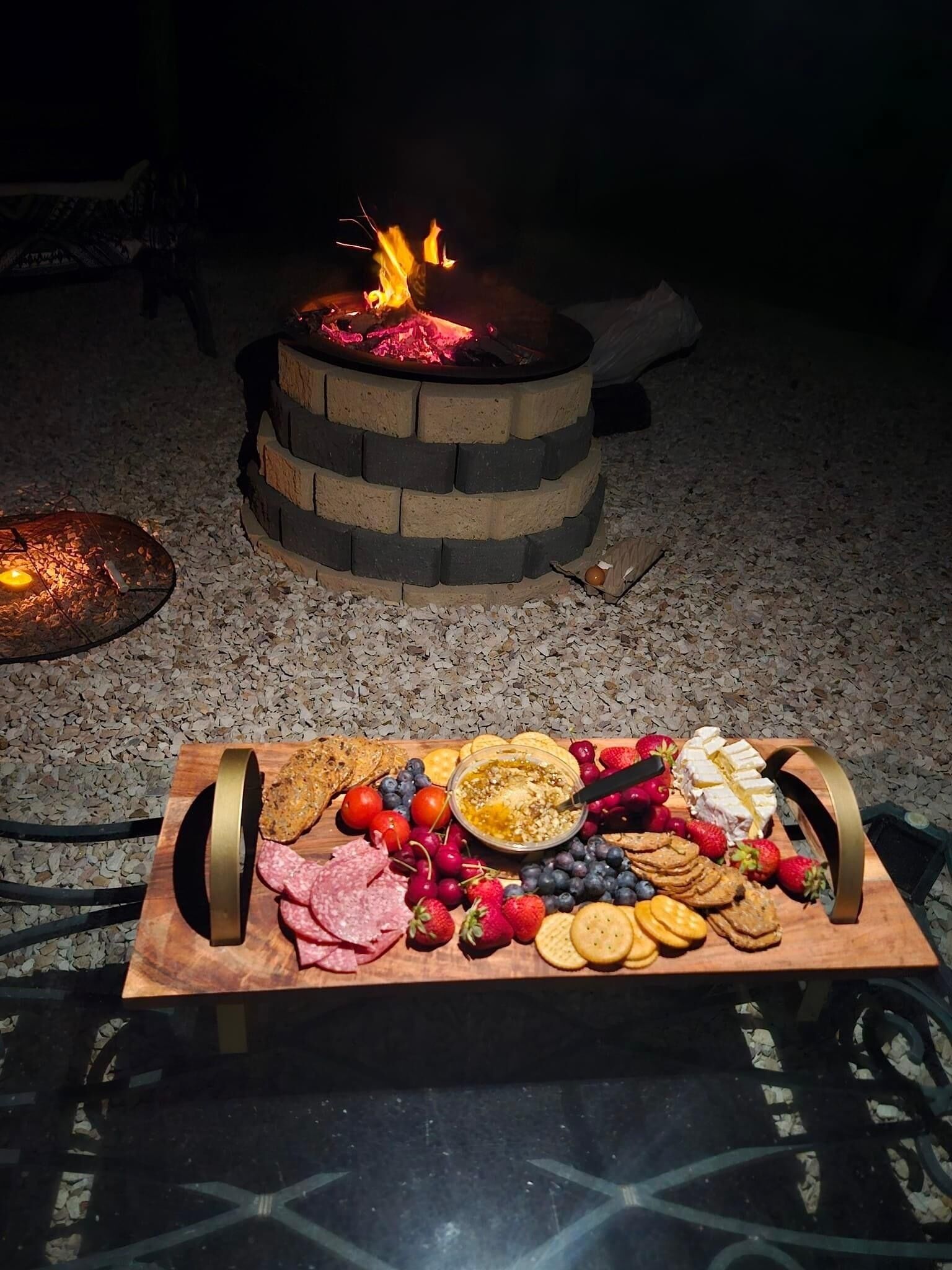Ein Lagerfeuer mit einem Feuerkorb aus Ziegelsteinen und einem Holzbord mit verschiedenen Snacks und Früchten.