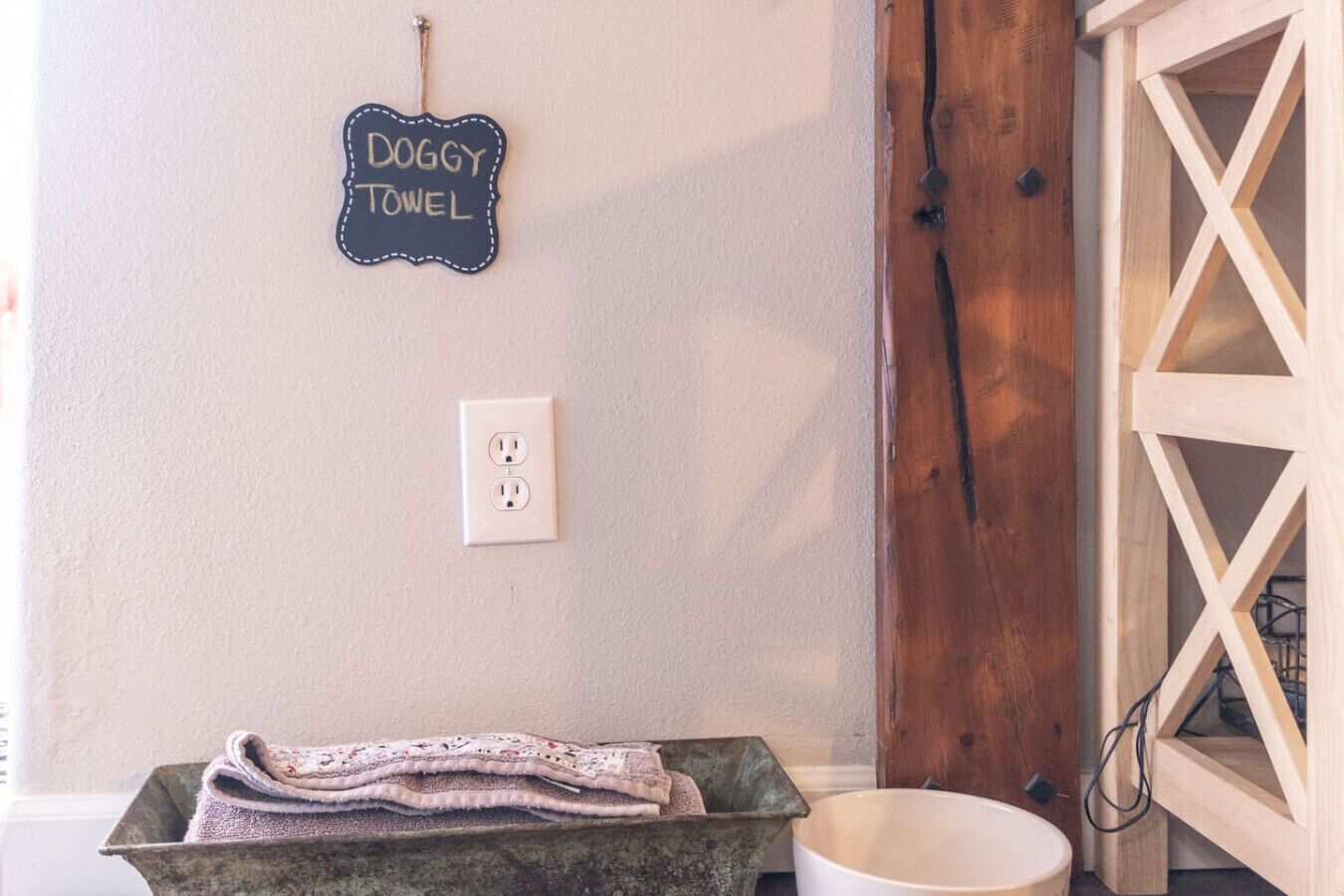 A wall-mounted sign reading "DOGGY TOWEL," a power outlet, a rustic wooden door, and a metal bucket with a towel.