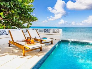 Un espace aménagé au bord de la piscine avec deux chaises longues, un plateau de fruits et un verre de jus d’orange, et une vue sur l’océan.