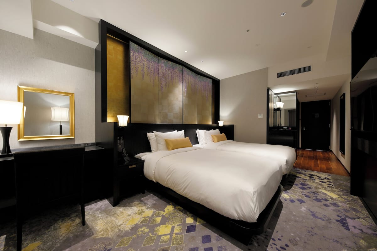 A modern hotel room with a large bed, a nightstand with a lamp, a wall-mounted mirror, and a decorative panel above the bed.