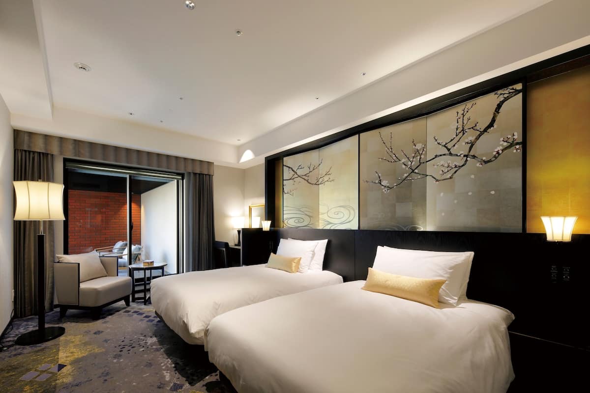A hotel room with two beds, a desk, a chair, and a large window with curtains.