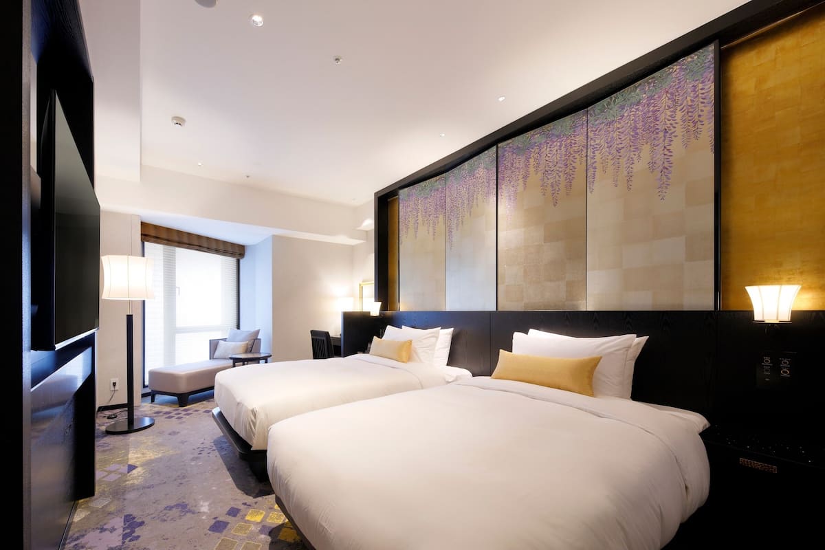 A hotel room with two beds, a desk, a chair, and a large artwork on the wall.