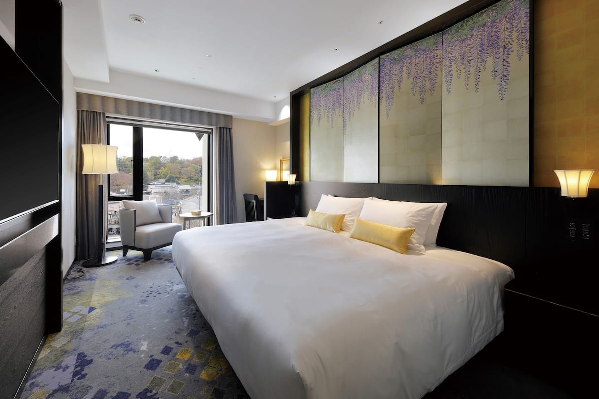 A hotel room with a large bed, a desk, a chair, a view through a window, and a decorative wall.