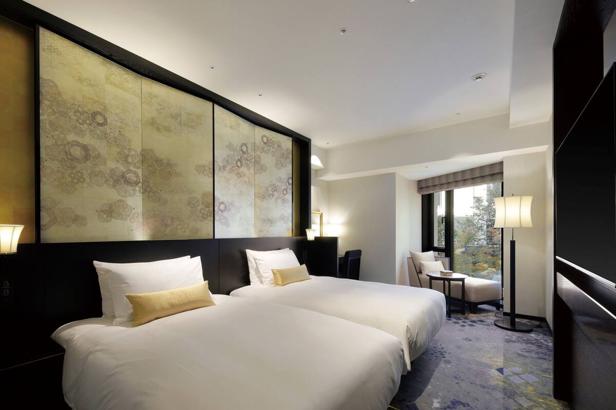 A hotel room with two beds, a large abstract artwork on the wall, a desk with a chair, and a view of the city through the window.