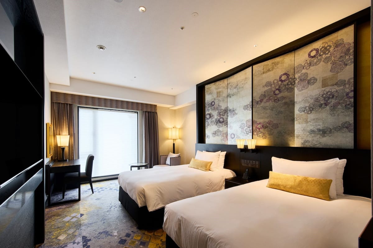 A hotel room with two beds, a desk, a chair, a large window, and a decorative wall piece.