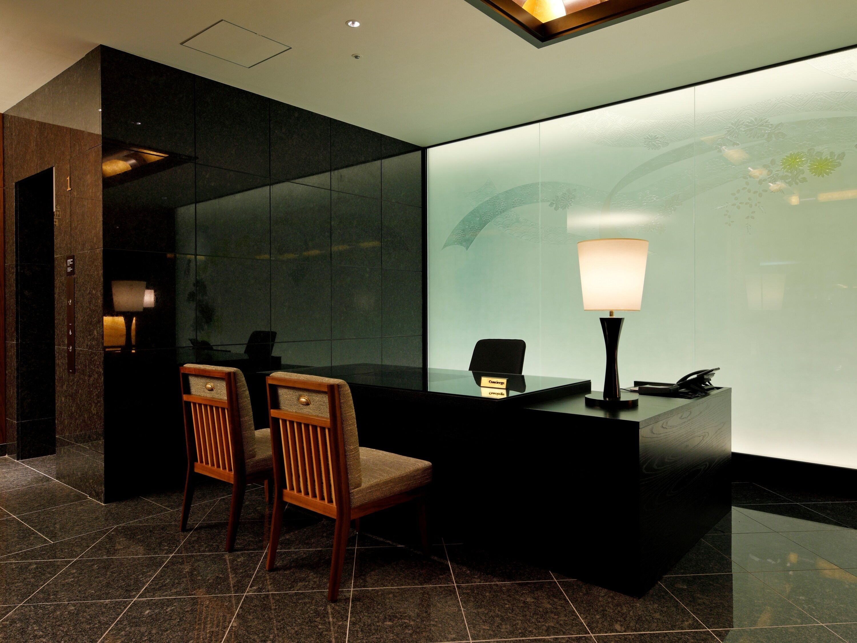 A reception desk with a glass partition, two wooden chairs, a lamp, and a telephone.
