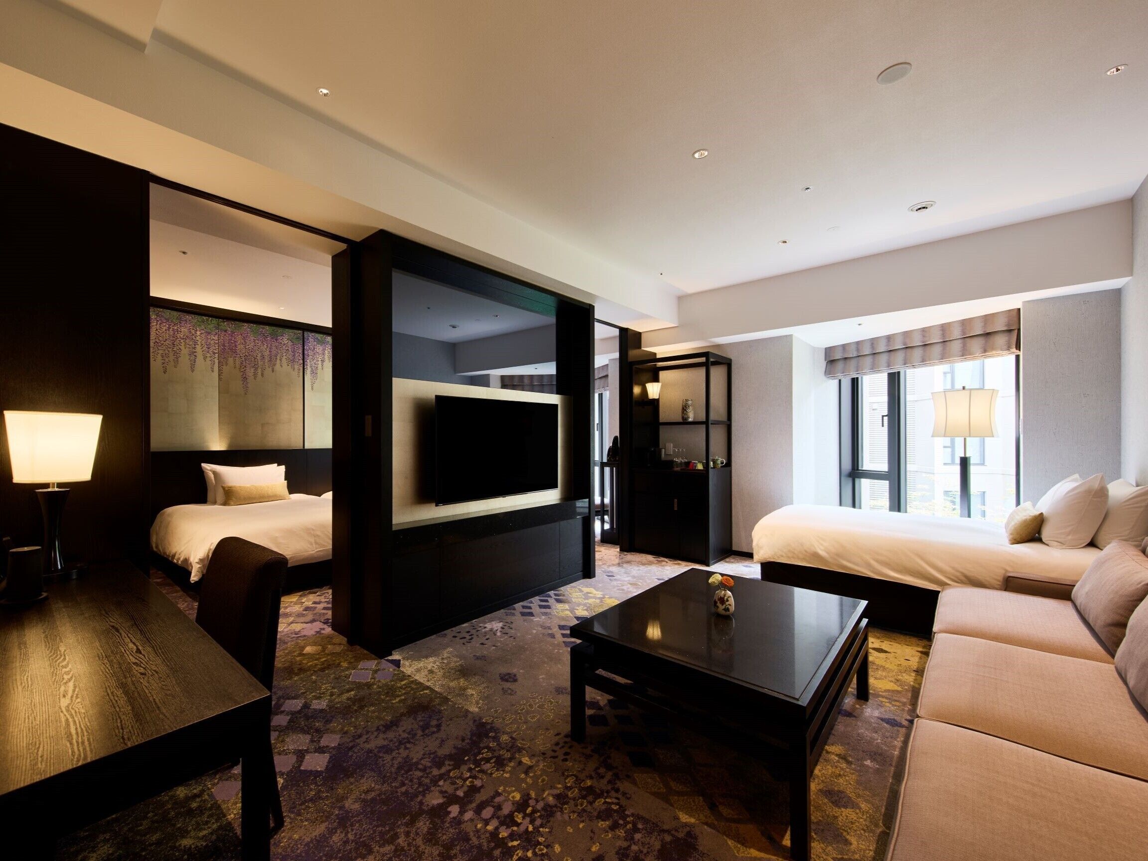 A hotel room with a bed, a sofa, a TV mounted on the wall, a coffee table, and a desk.