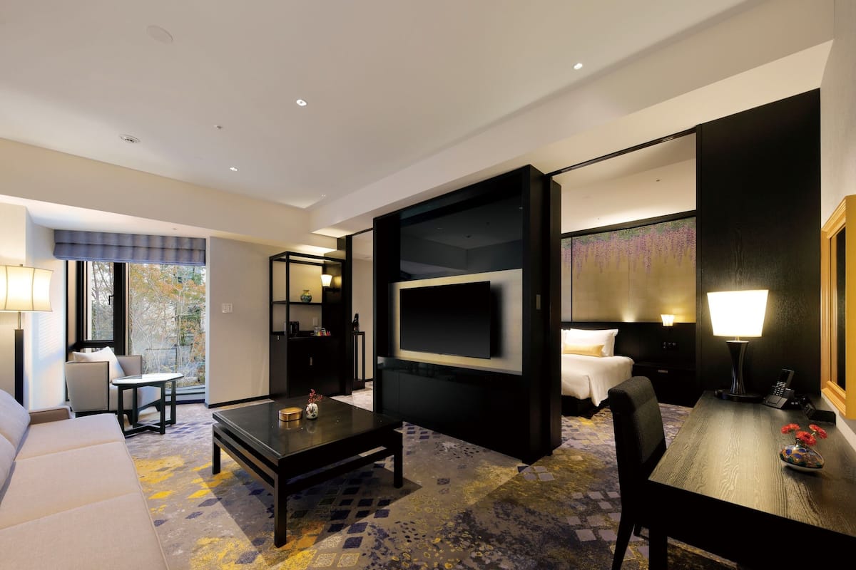 A modern hotel room with a sofa, a coffee table, a TV mounted on the wall, a desk with a lamp, and a bed with a nightstand.
