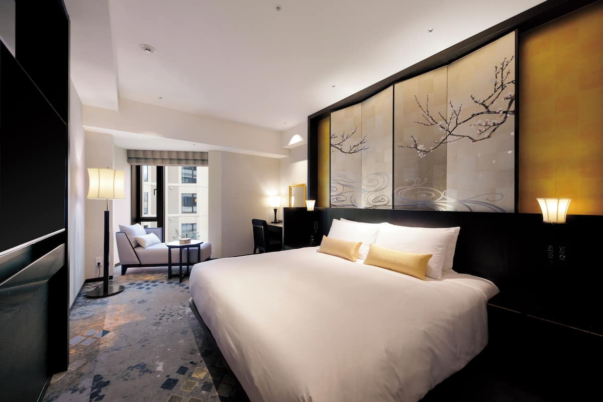 A hotel room with a large bed, a desk, a chair, and a view of the city through the window.