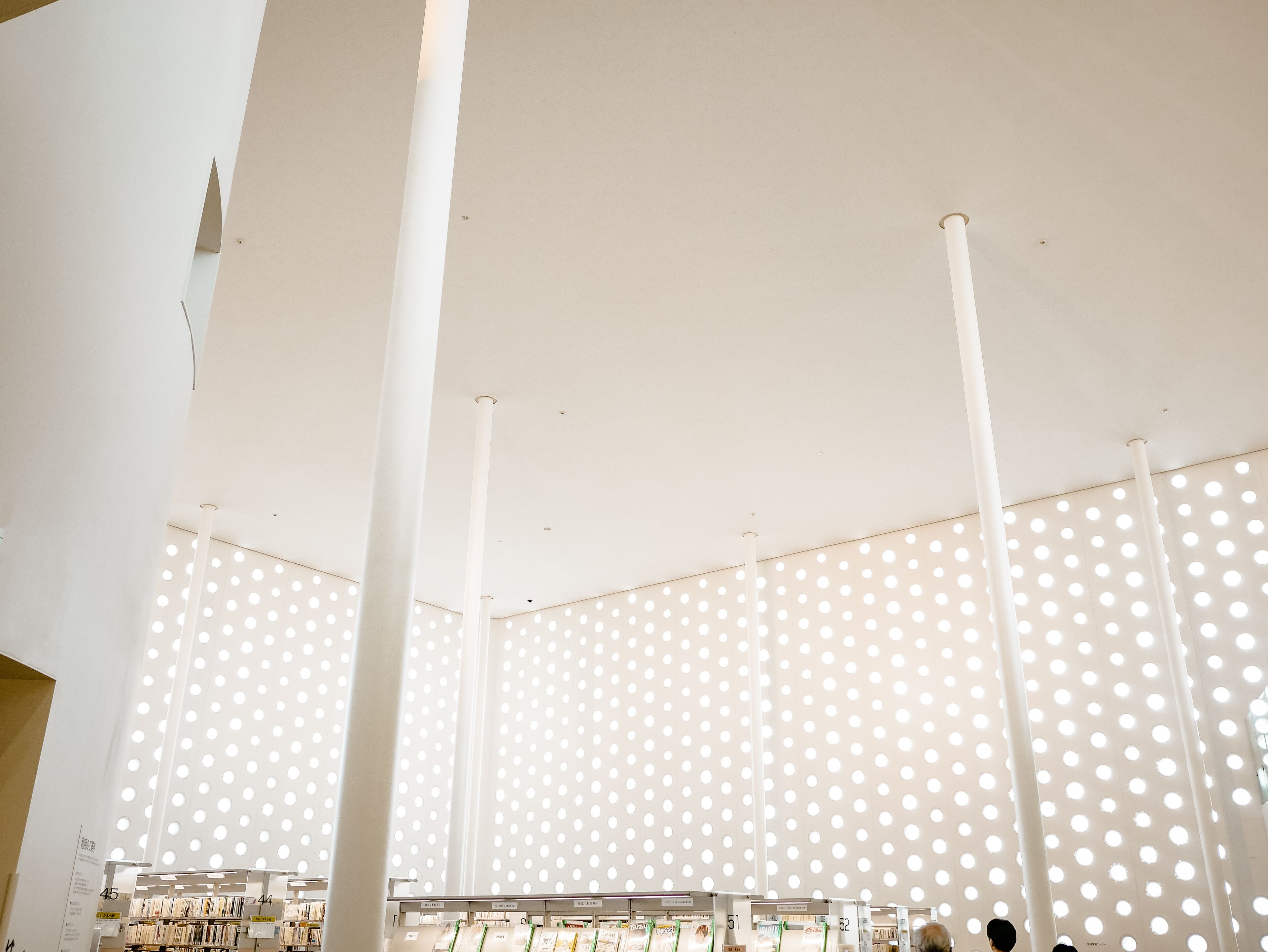 Un intérieur moderne avec des colonnes blanches et un plafond orné d’ouvertures circulaires. On aperçoit des étagères remplies de livres au premier plan.