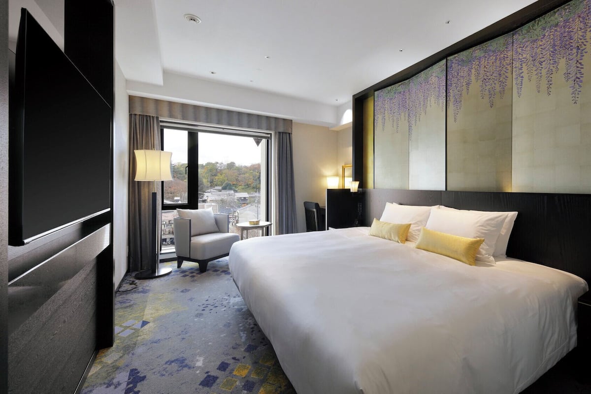 A hotel room with a large bed, a desk, a chair, a TV, and a view of the city.