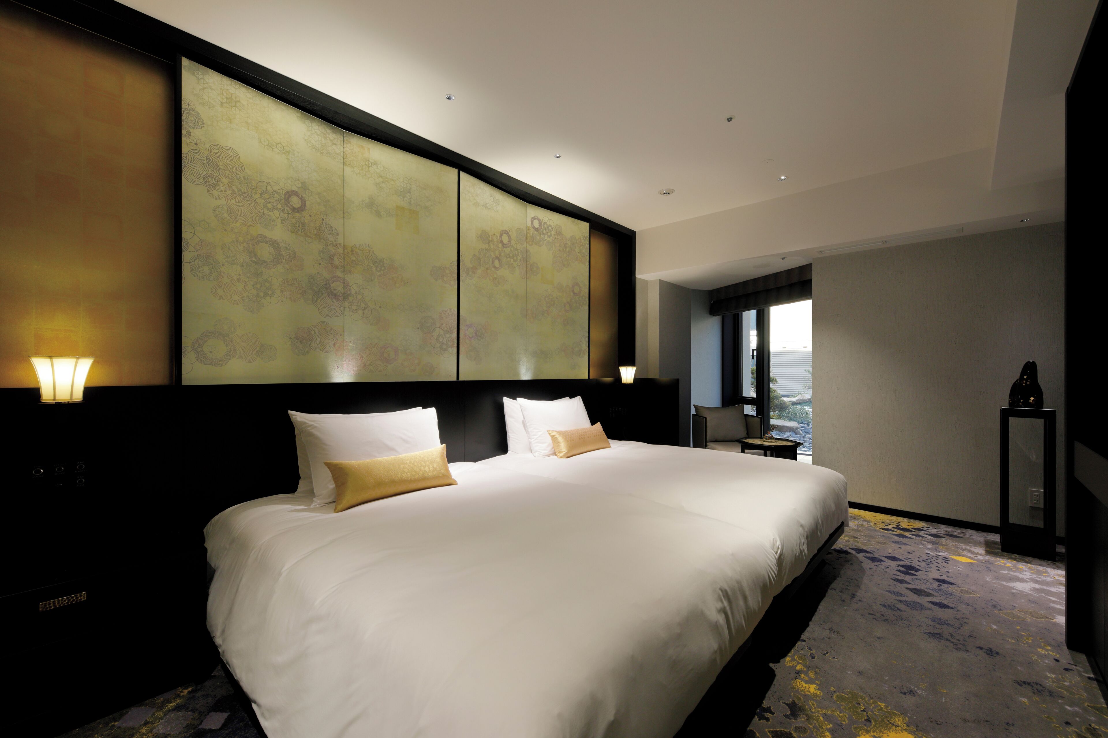 A modern hotel room with a large bed, a headboard with decorative panels, a nightstand, and a view of the outdoors.