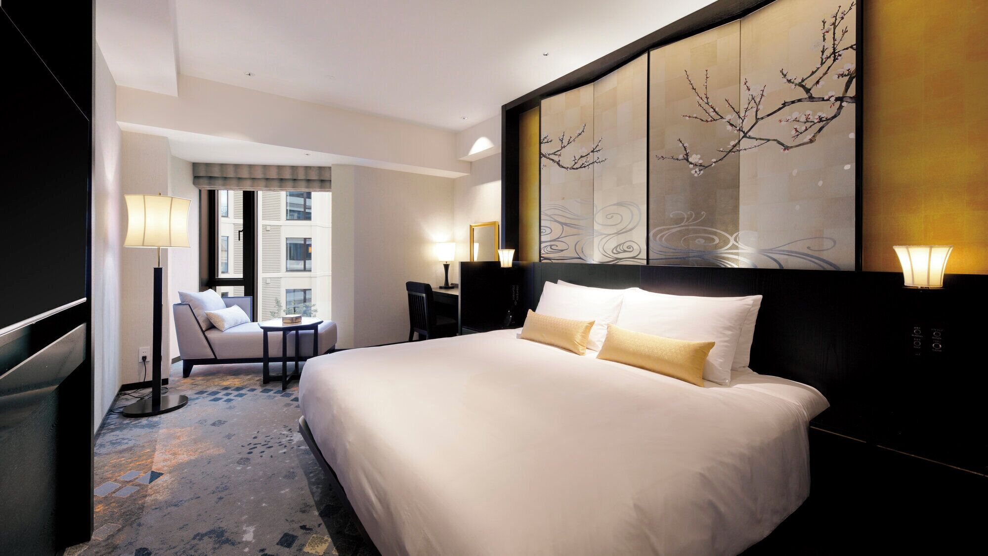 Quarto de hotel com uma cama grande, uma escrivaninha, uma cadeira e vista para a cidade pela janela.