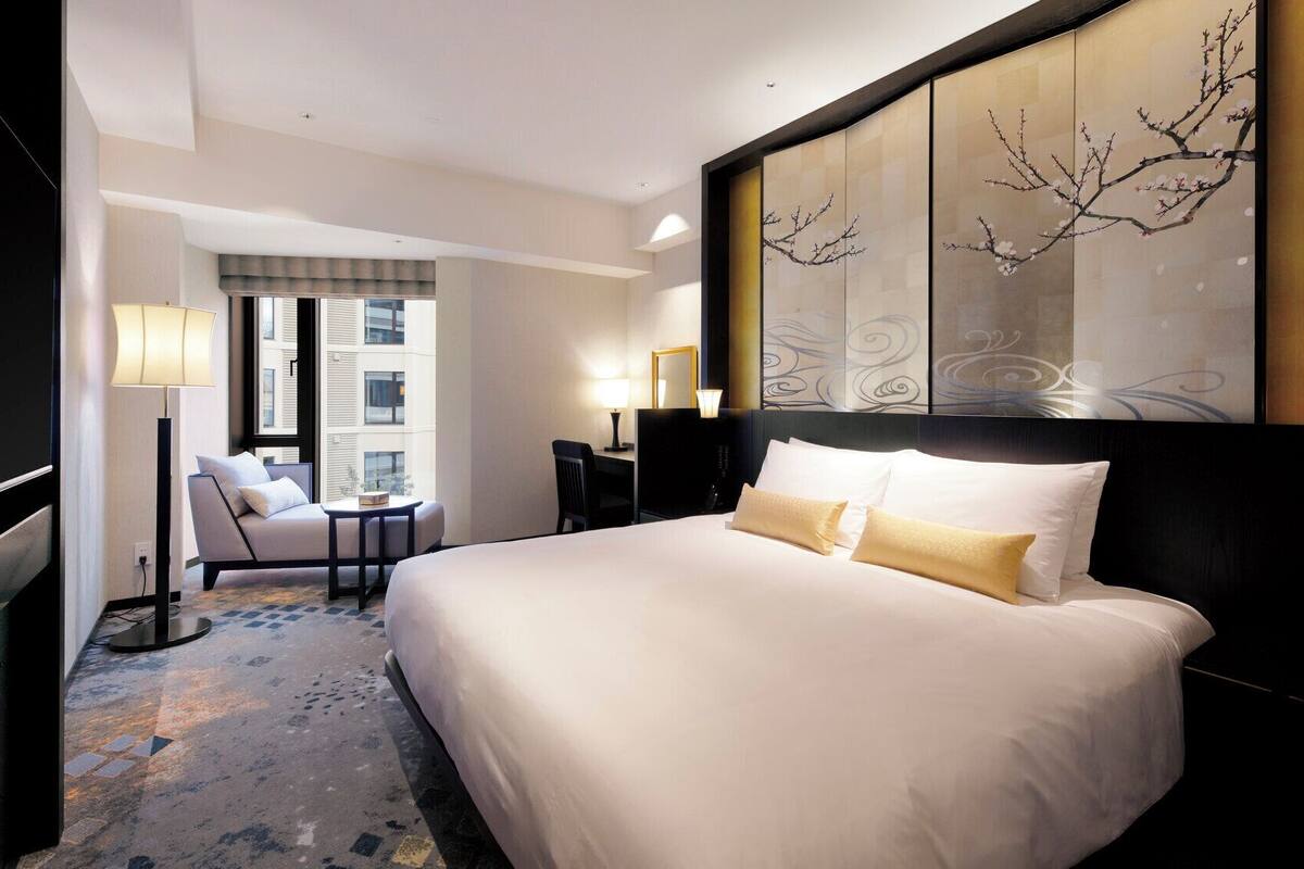 A hotel room with a large bed, a desk, a chair, and a view of the city through the window.