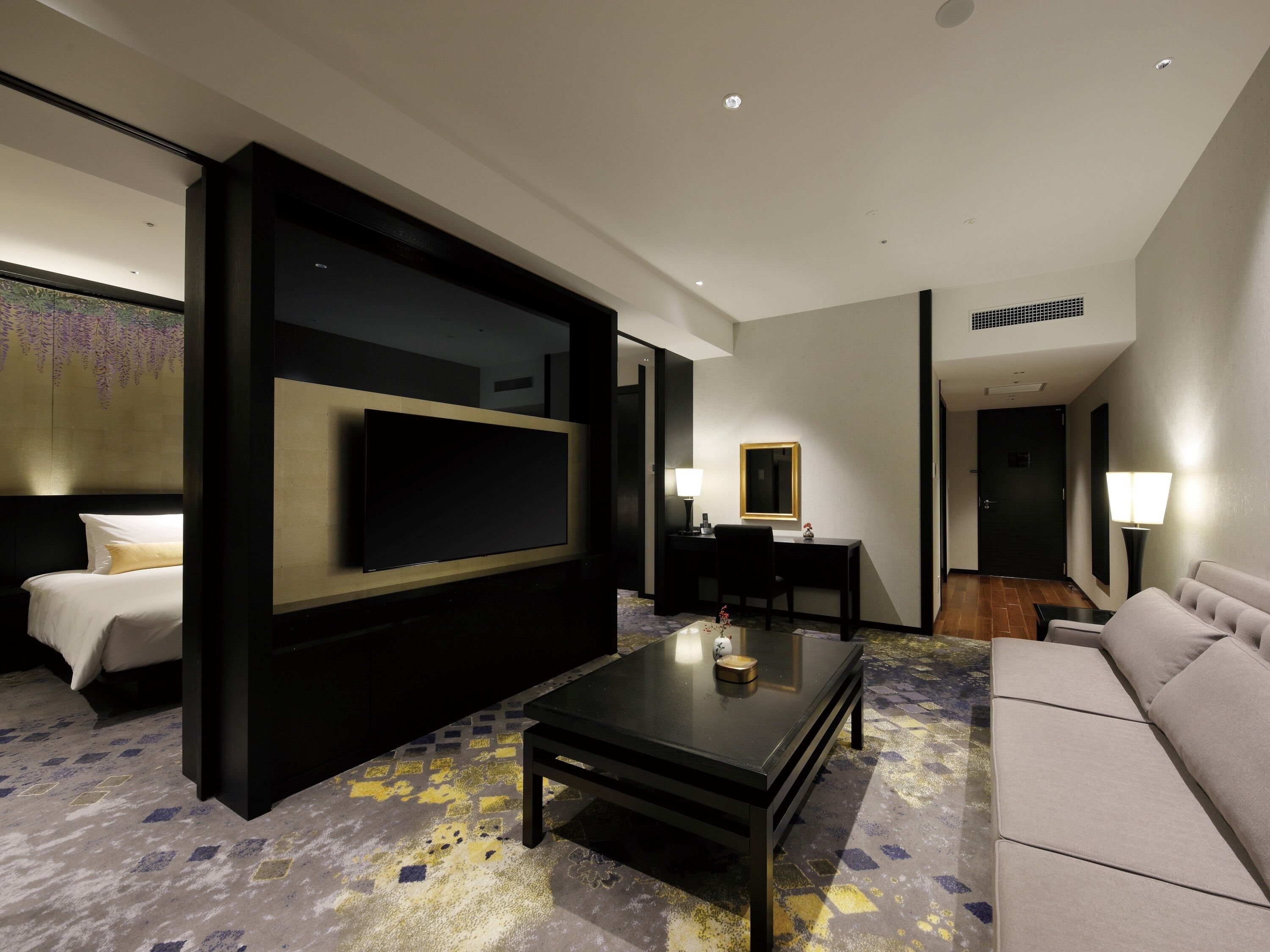 Une chambre d’hôtel moderne équipée d’un grand téléviseur à écran plat, d’un canapé, d’une table basse et d’un bureau avec une lampe.