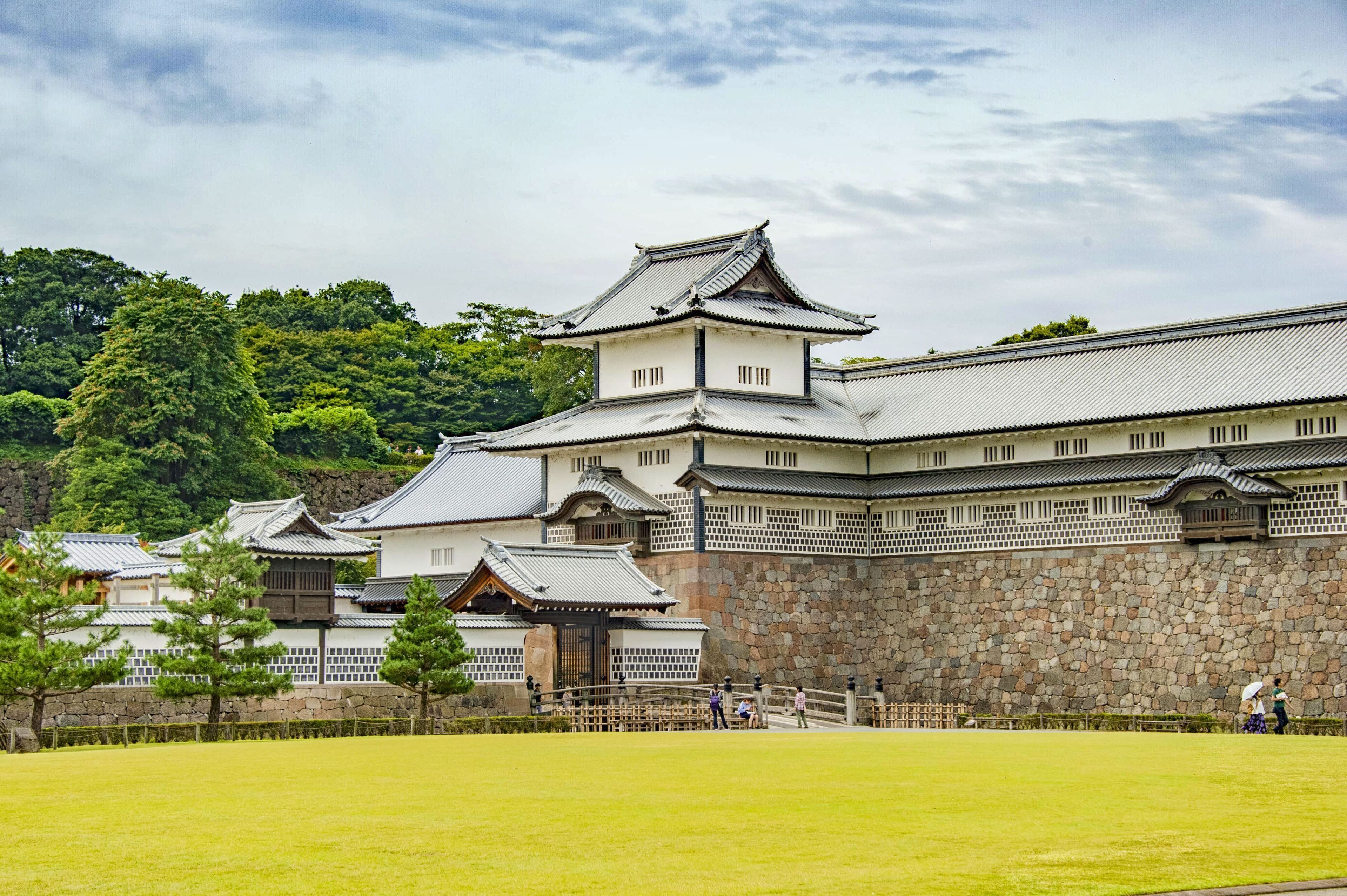 Un château japonais traditionnel composé de plusieurs bâtiments, d’un mur de pierre et d’une cour verdoyante.