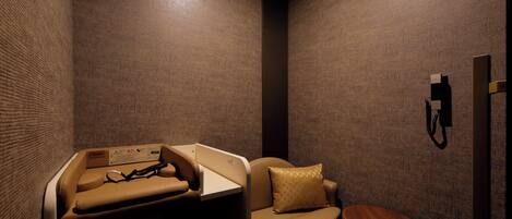 A small, dimly lit room with a massage table, a cushioned chair, and a small wooden table.