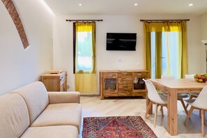 A living room with a sofa, a dining table with chairs, a TV mounted on the wall, and a wooden cabinet with drawers.