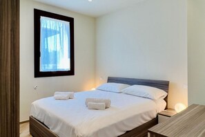 A neatly made bed with white linens and two towels, a bedside table with a lamp, and a window with curtains.