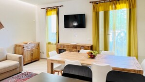 Un saló amb un sofà, una taula de dinar amb cadires, un televisor penjat a la paret i una finestra amb cortines.