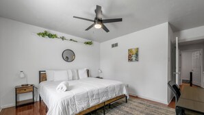 A bedroom with a bed, a ceiling fan, a bedside table, a nightstand, a wall-mounted mirror, a small shelf with plants, and a door leading to another room. - Your Kentucky Home (Georgetown)