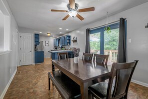 A dining area with a wooden table and chairs, a ceiling fan, and a view of the outdoors through large windows.
