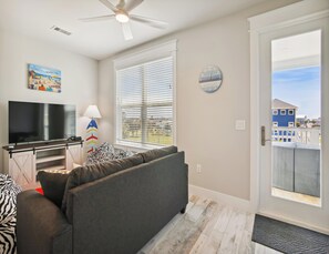 A living room with a flat-screen TV, a ceiling fan, a large window with blinds, a clock, and a view of a blue house through the window. - Pearl of the Sea by Ryson Vacation Rentals (Galveston)