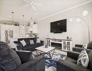 Ein modernes Wohnzimmer mit einer grauen Sofagarnitur, einem Couchtisch und einem Kamin. Es gibt eine Küchenzeile mit weißen Schränken und einem flachbildfernseher, der über der Arbeitsfläche montiert ist.