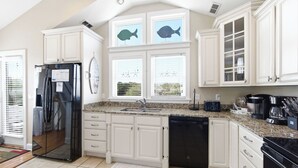 Una cocina con armarios blancos, un refrigerador negro, lavavajillas, ventana con vistas y decoración con una tabla de surf.