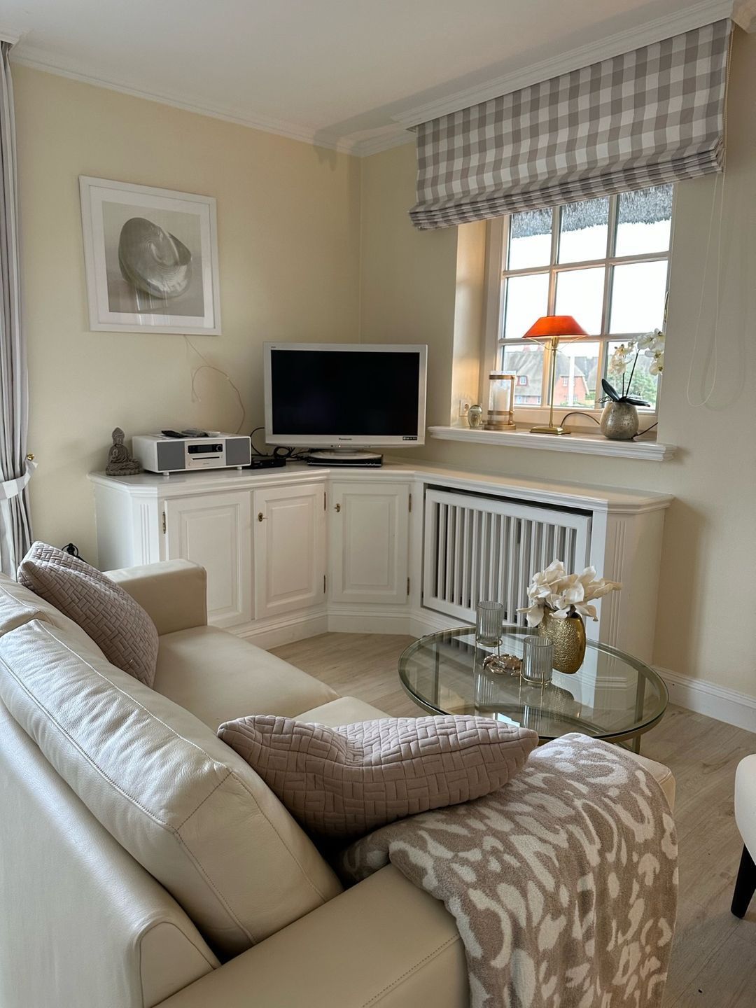A living room with a beige sofa, a glass coffee table, a flat-screen TV, and a window with a checkered valance.