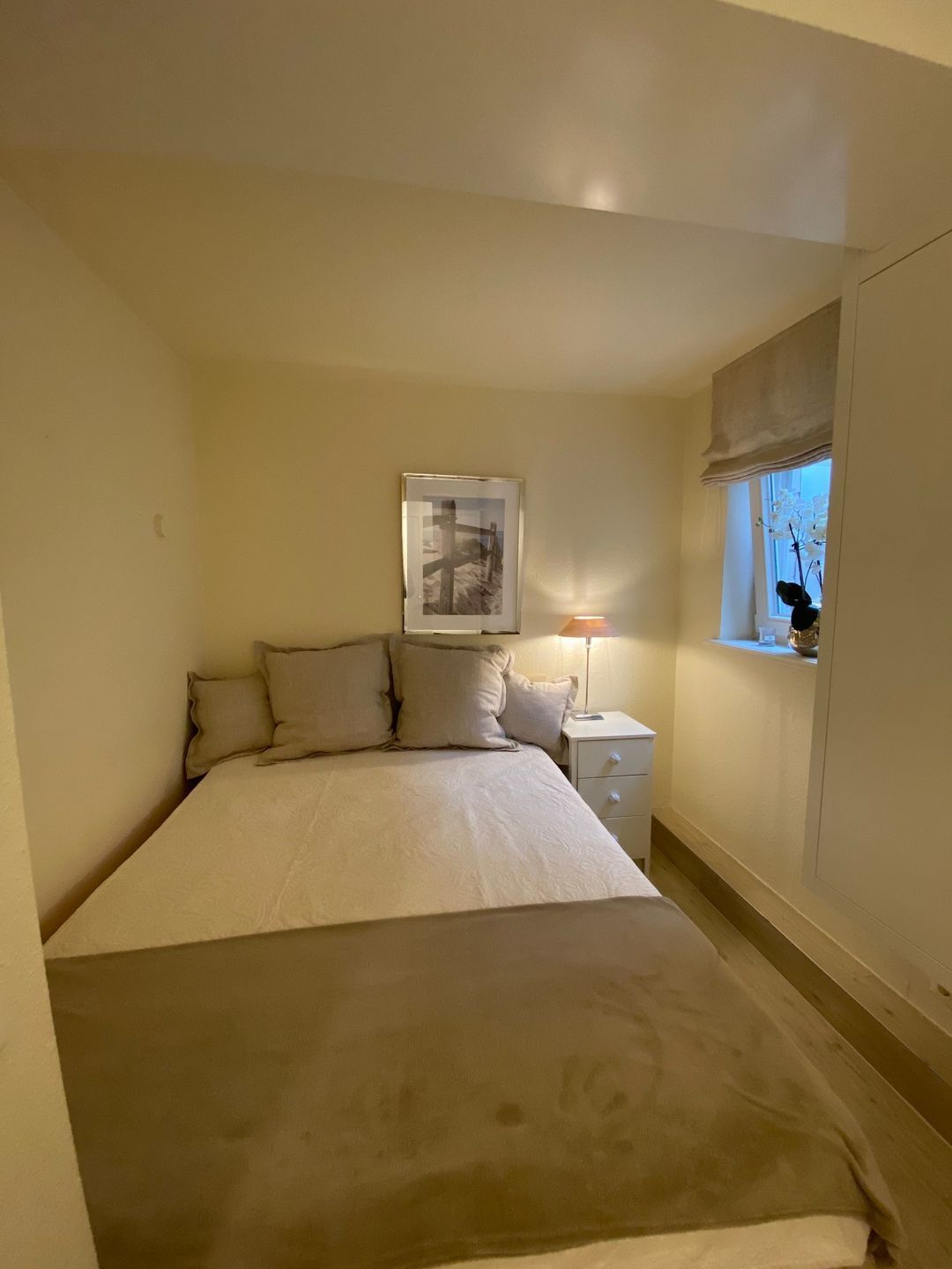 A bedroom with a bed, a nightstand, a lamp, a window with a view, and a framed picture on the wall.