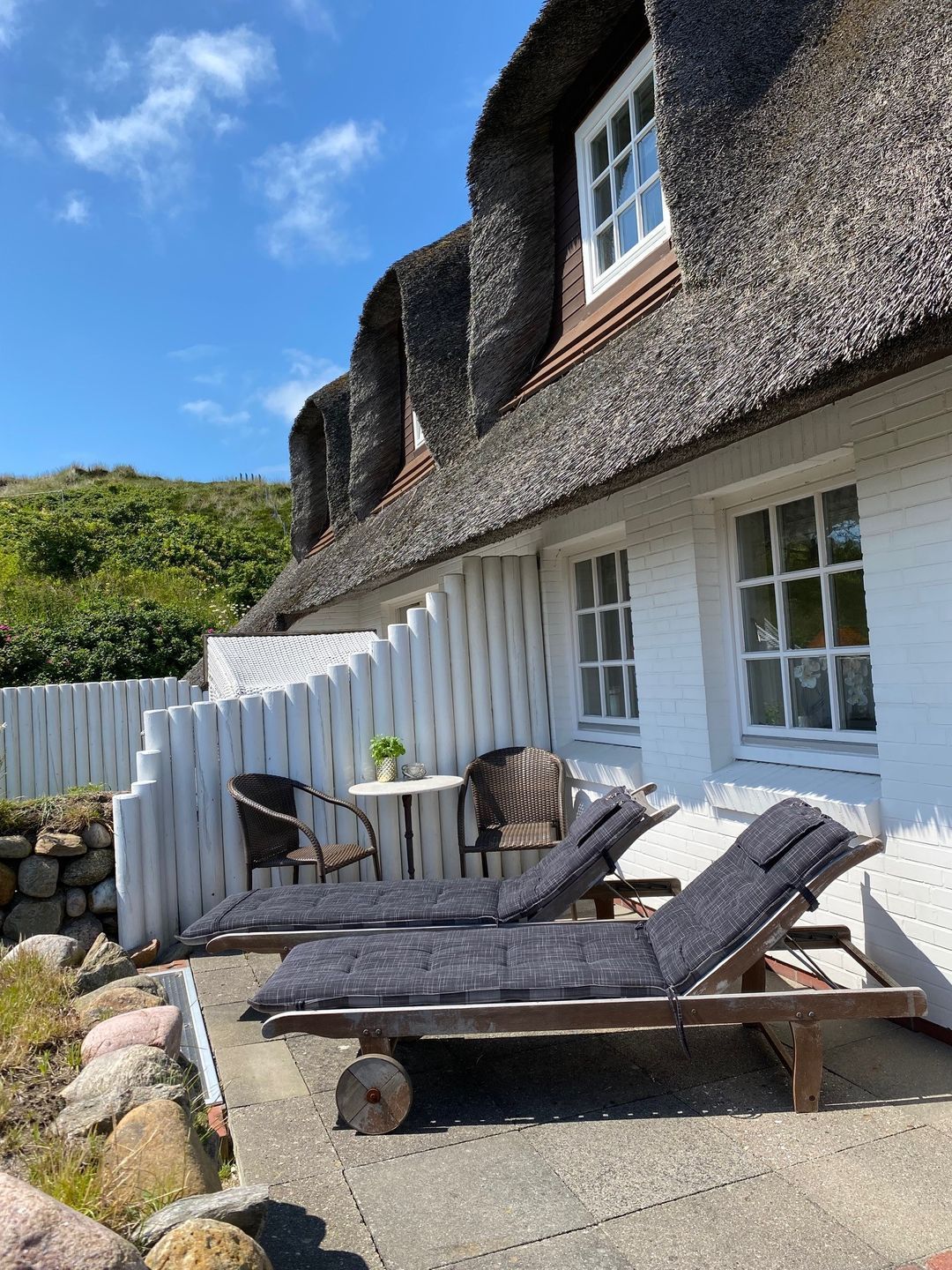 A thatched-roof house with a white exterior, a small patio with lounge chairs, and a stone wall.