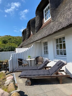 A thatched-roof house with a white exterior, a small patio with lounge chairs, and a stone wall. - Haus Wattblick App. 06 - Wattblick (Sylt)