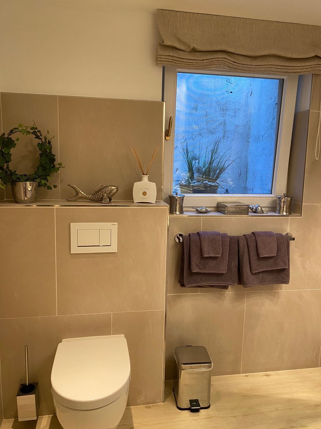 Une salle de bain comprenant des toilettes, une poubelle, une fenêtre avec vue et une étagère sur laquelle se trouvent un pot de fleurs et un objet décoratif.
