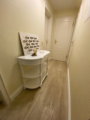 A narrow hallway with a white door at the end, a white wall with a decorative panel, and a white storage unit with shelves. - Haus Wattblick App. 06 - Wattblick (Sylt)