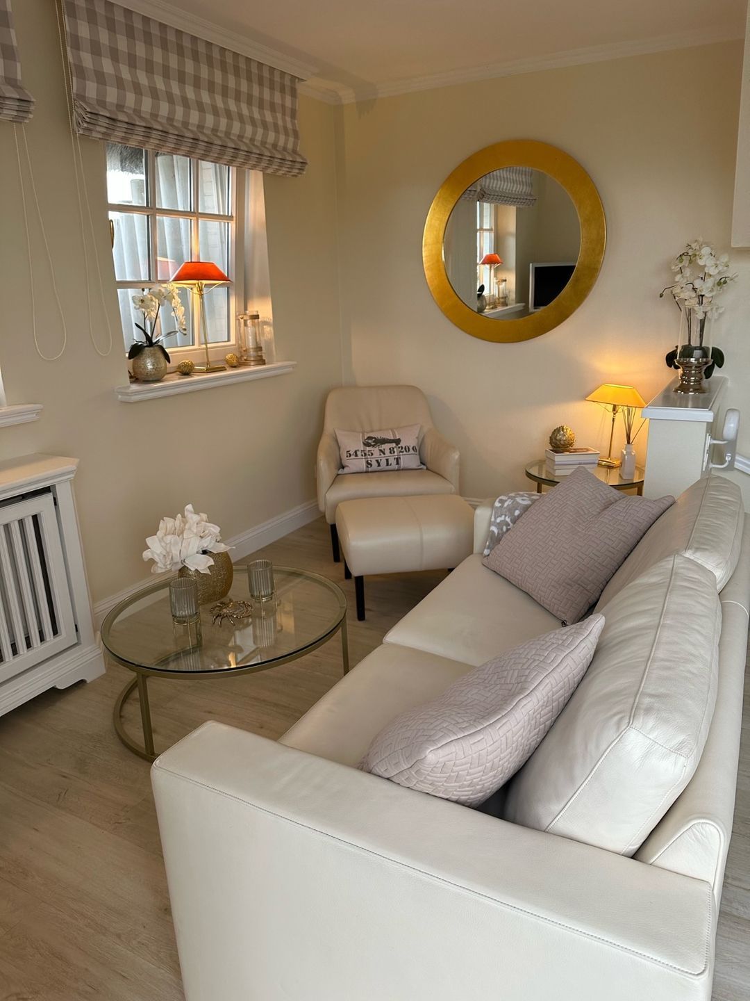 A living room with a white leather sofa, a round coffee table, a chair, a round mirror, and a window with curtains.