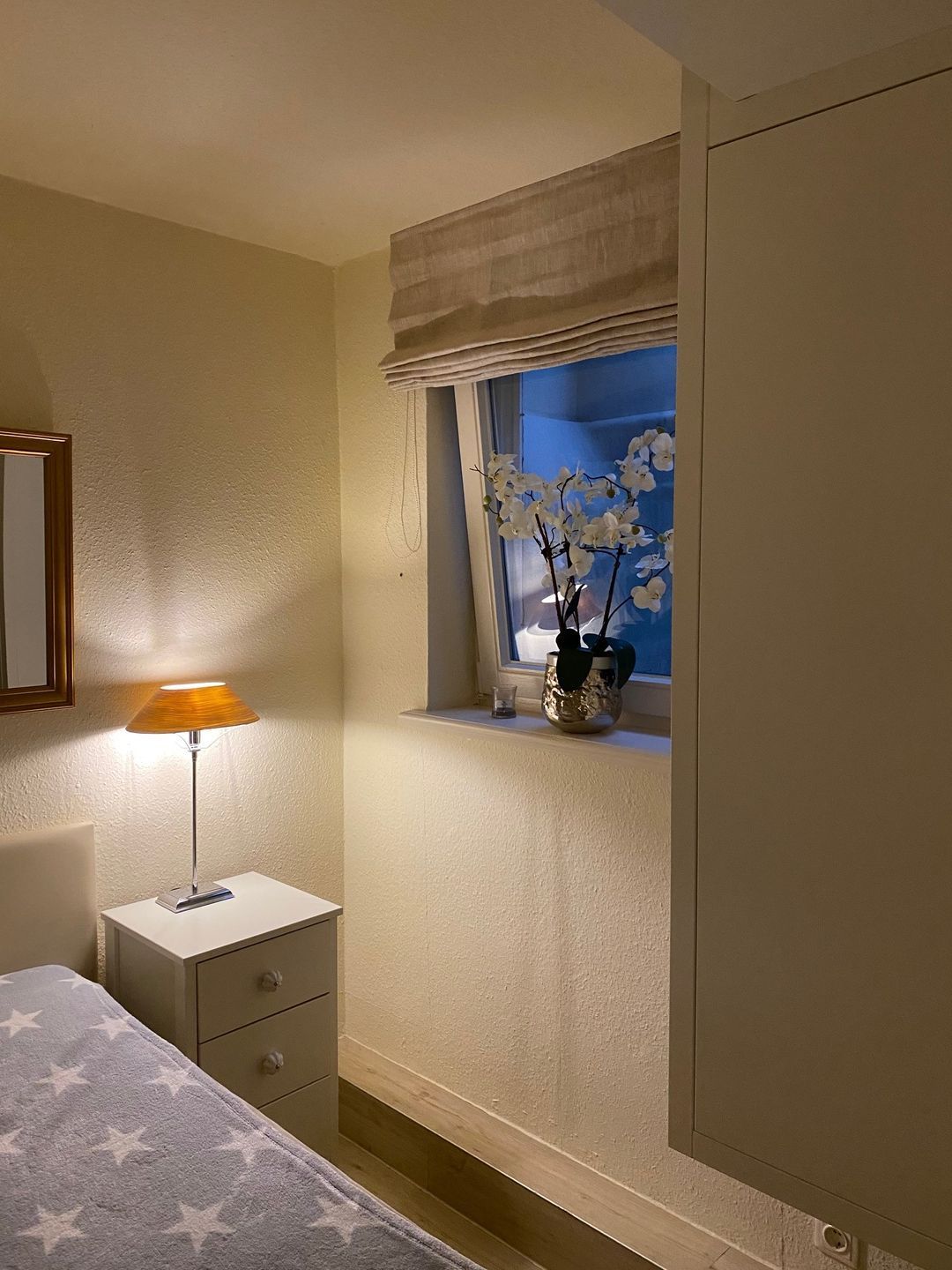 A bedroom with a bed, a bedside table with a lamp, a window with a view of white flowers, and a cabinet.