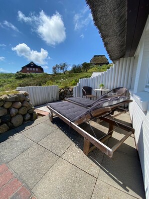 Une terrasse avec deux chaises longues, un mur en pierre et une structure surmontée d’un toit de chaume.