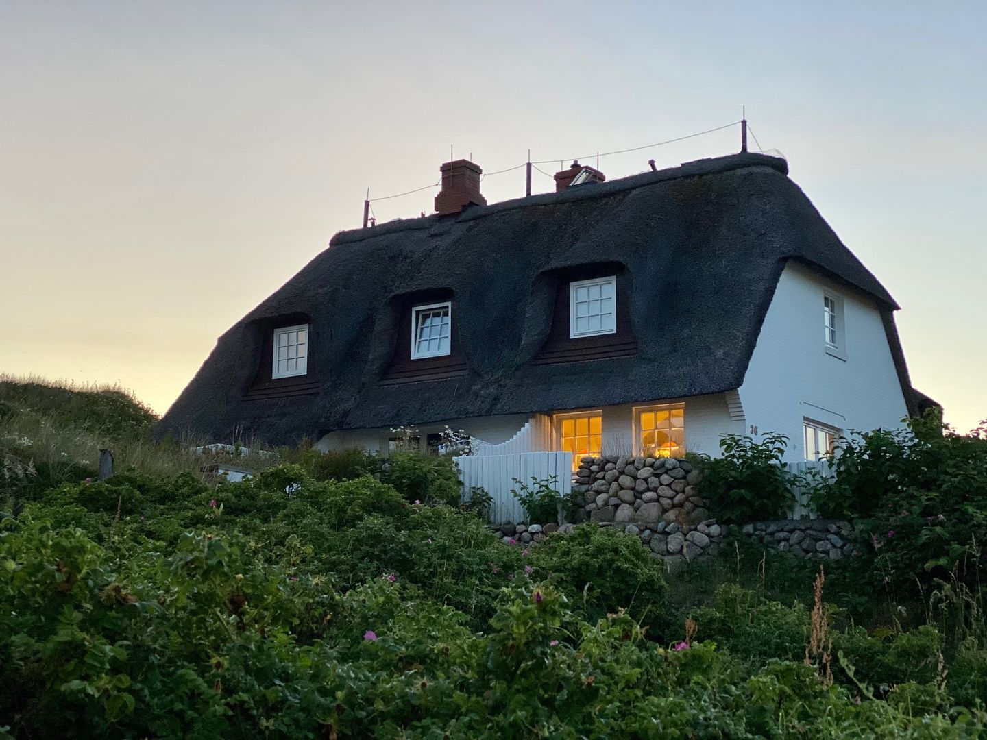 Ein traditionelles Reetdachhaus mit drei Fenstern und einer Mauer aus Stein davor.
