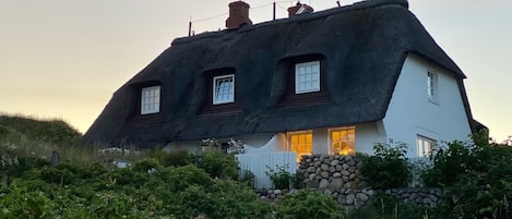 A traditional thatched-roof house with three windows and a stone wall in front.