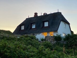 Une maison traditionnelle à toit de chaume, avec trois fenêtres et un mur de pierre à l’avant.