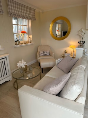 Un salon avec un canapé en cuir blanc, une table basse ronde, un fauteuil, un miroir rond et une fenêtre avec des rideaux.